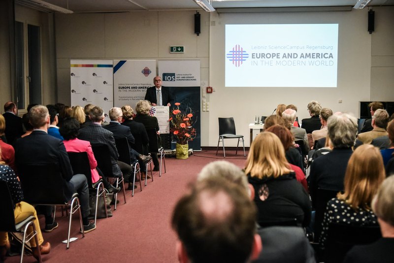 Matthias Kleiner, Präsident der Leibniz-Gemeinschaft, hält ein Grußwort. Hinter ihm ist das Logo des WissenschaftsCampus an die Wand projiziert. Die Gäste sind von hinten zu sehen.. Foto: Julia Dragan